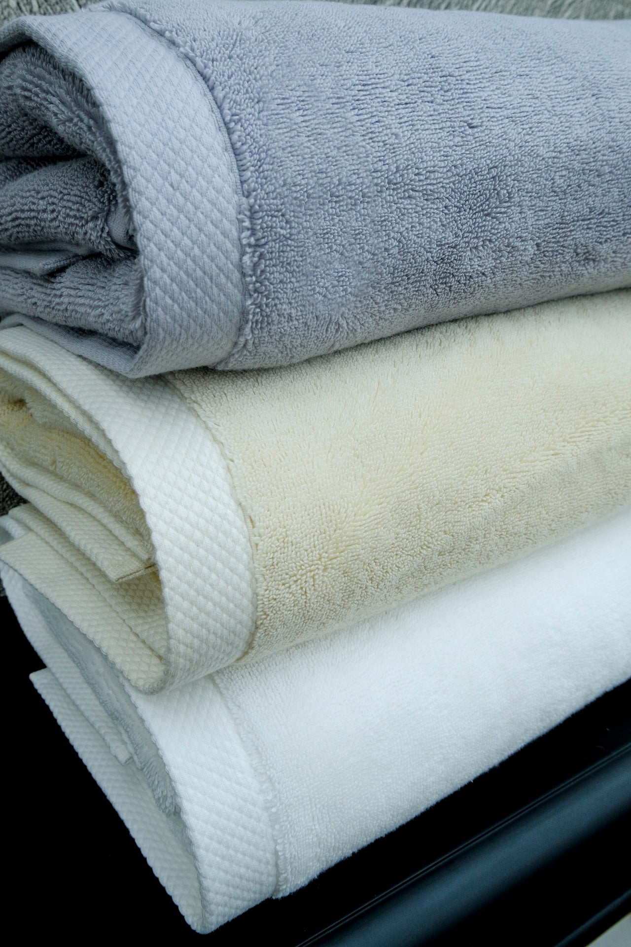 Close-up of a stack of three luxury organic cotton bath towels in white, cream, and grey from Minoa Home, displayed on a marble background