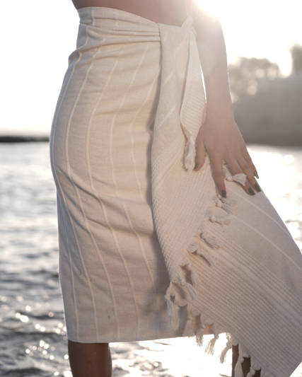 Person wearing a white towel with fringes by a body of water.