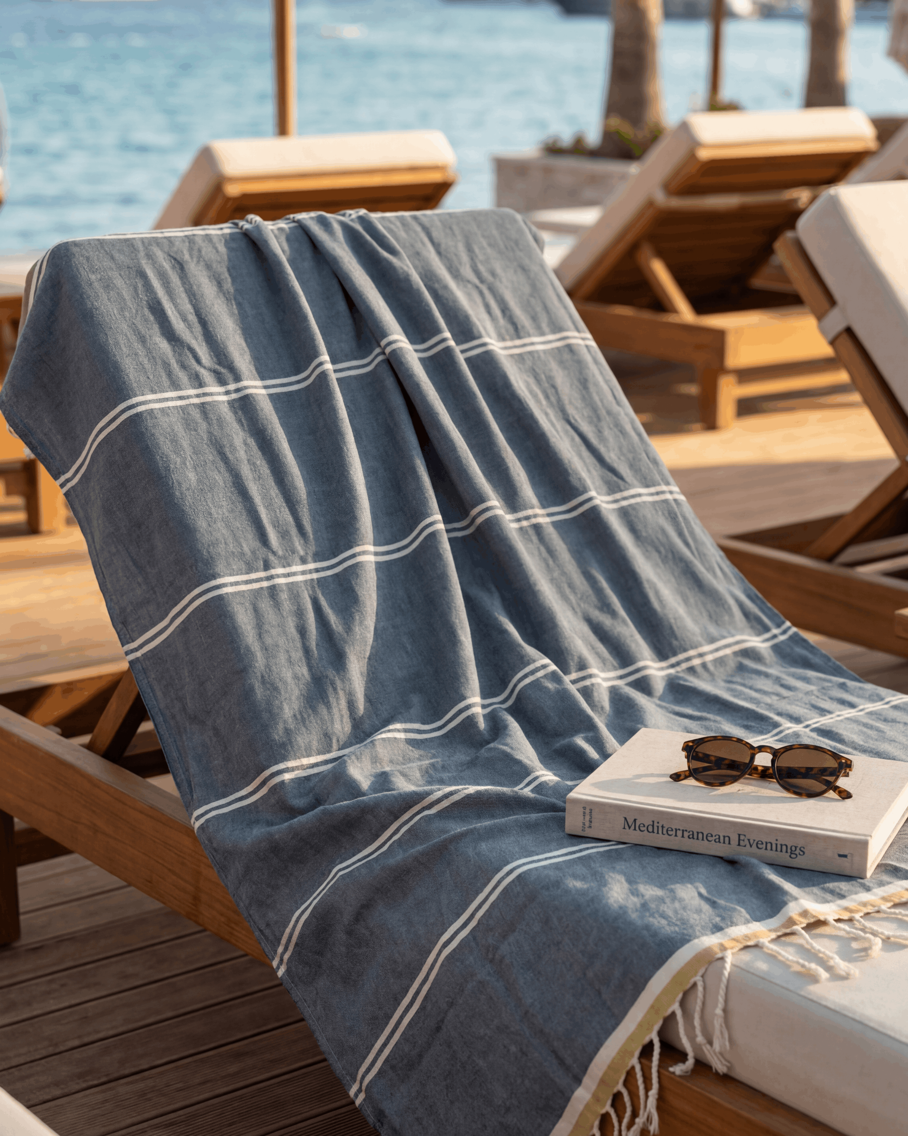 A blue and white striped cotton beach towel with fringe trim draped over a wooden lounge chair on a sunny beach deck, with the ocean and palm trees in the background.