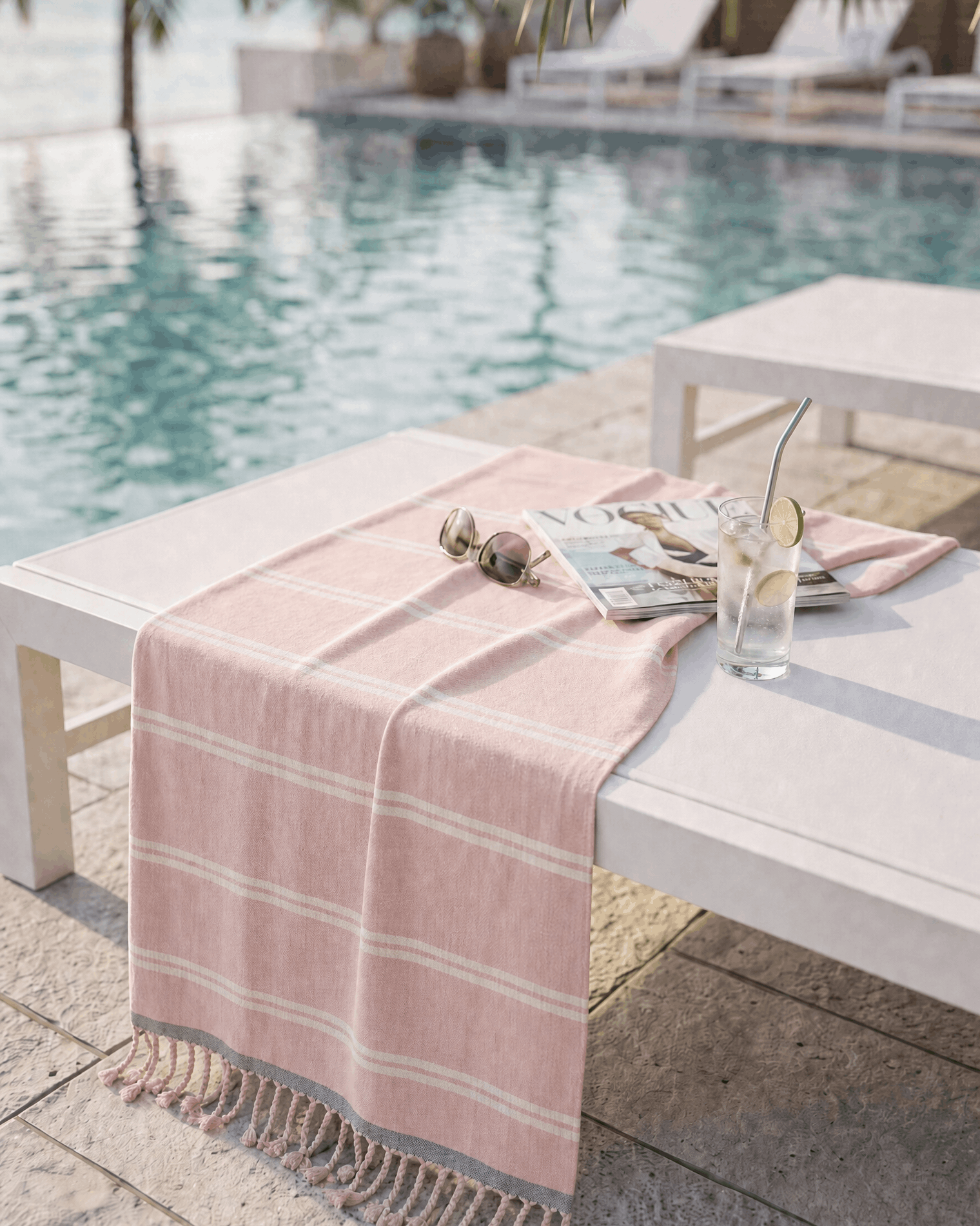 A pink and white striped cotton beach towel with fringed edges draped over a white outdoor lounge chair next to a swimming pool with palm trees in the background.