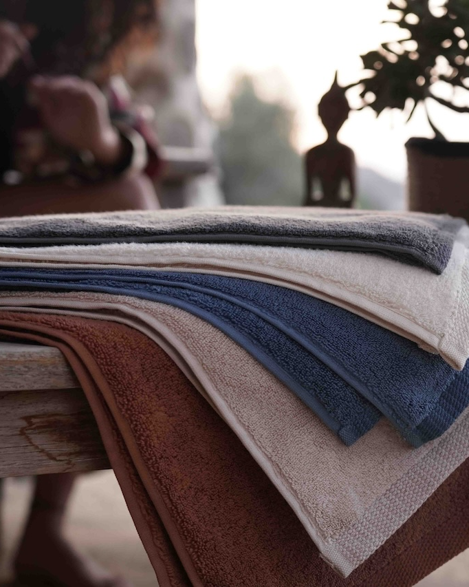 Plush Lite 500 gram Aegean Cotton large bath and hand towel set in Moonlight Pearl Honey Latte Charcoal Embrace Blue Velvet Cinnamon Charm and Pure n Raw arranged on a tree pattern table beside a woman showcasing sustainable luxury with soft organic bath towels and luxury bath towels