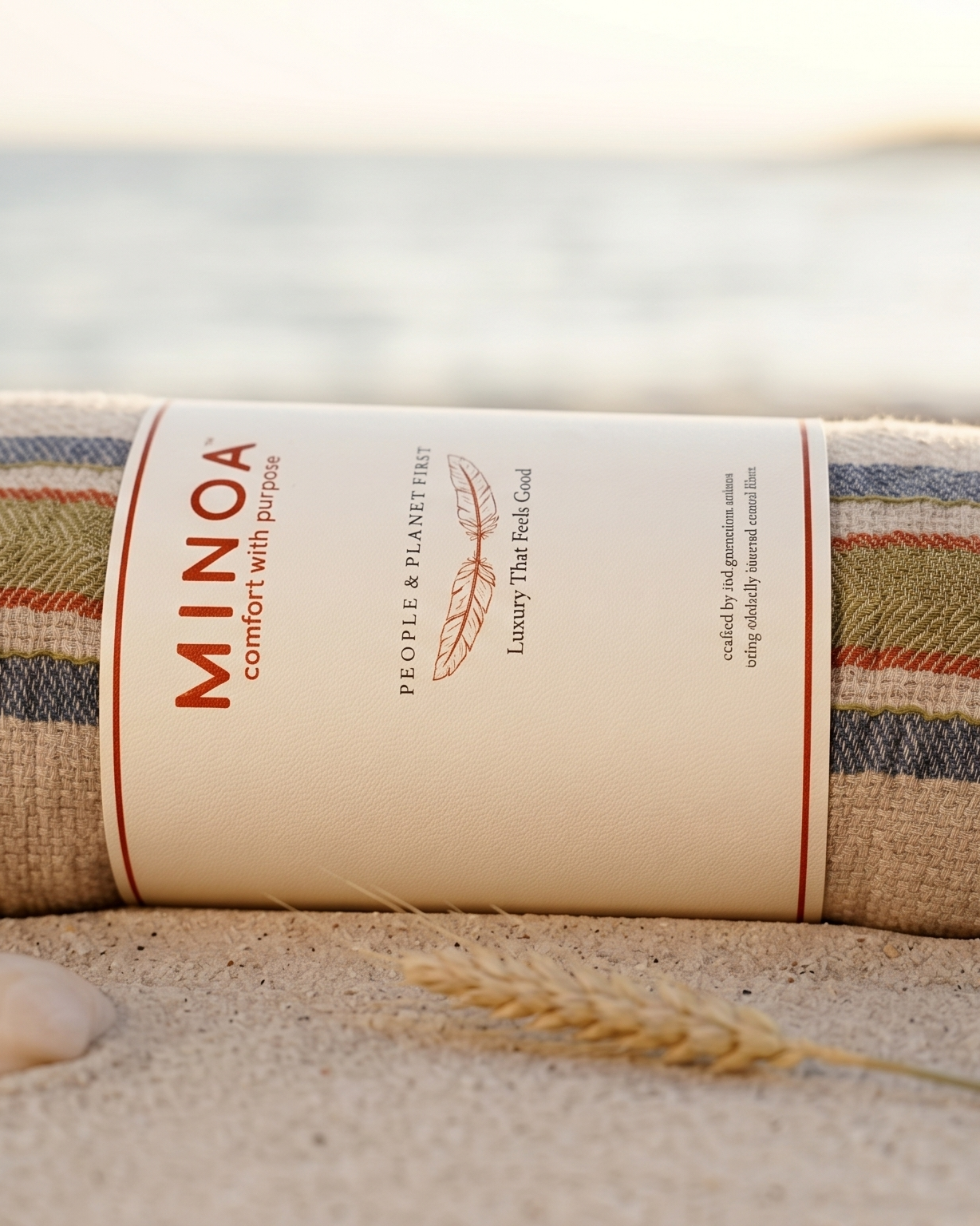 A rolled beach towel with a cream paper label reading MINOA, placed on sandy ground with shells and a blurred ocean background at sunset.