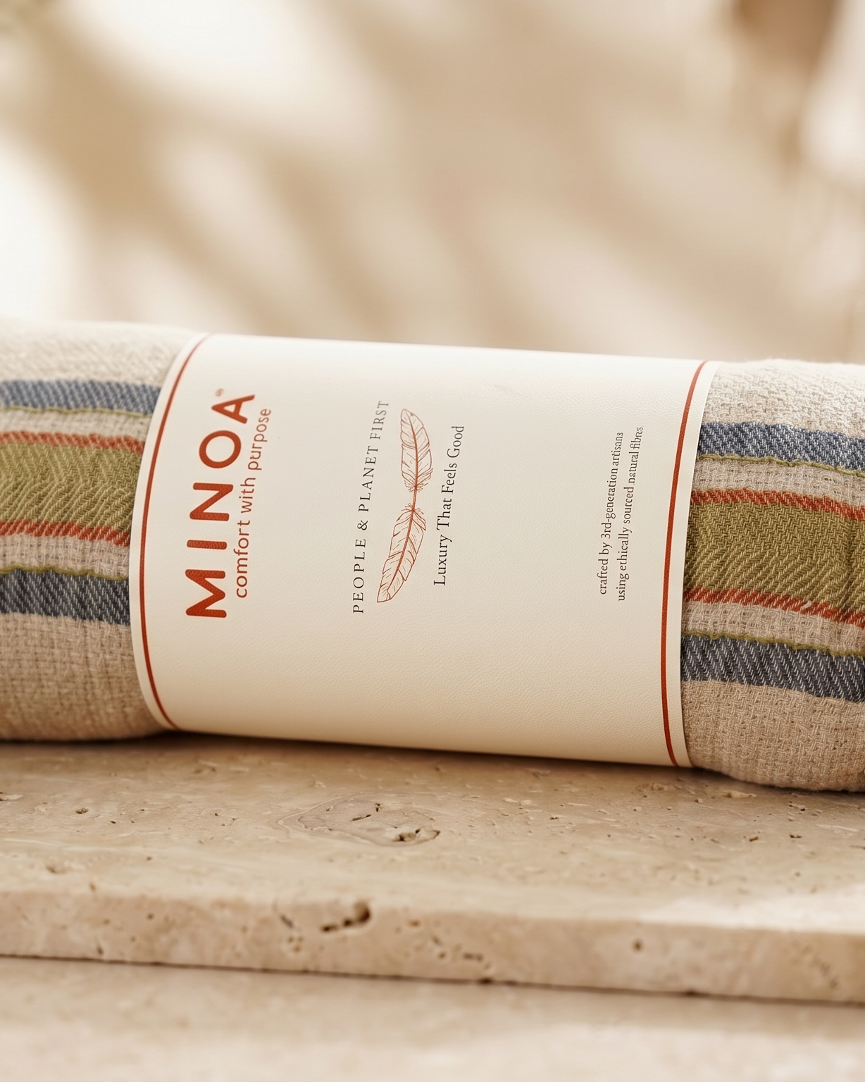 A rolled linen towel with a cream paper band labeled MINOA, placed on a stone surface with dried lavender, shells, and soft neutral props in the background.