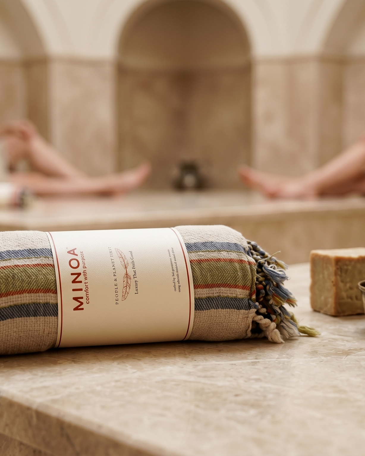 A striped spa towel wrapped in a paper band labeled MINOA, displayed on a stone table in a warm, blurred spa interior with two women relaxing in the background.