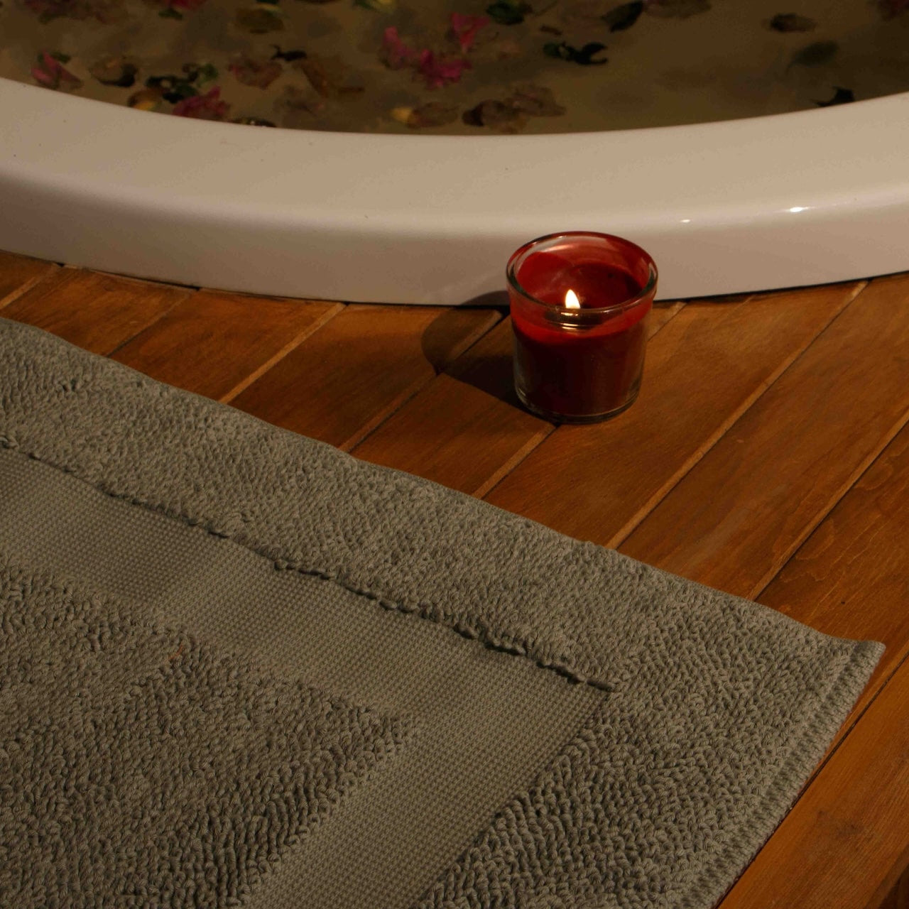 Candle in a red holder on a wooden surface next to a bathtub with a Minoa's Kaia Mat showcasing spa feeling