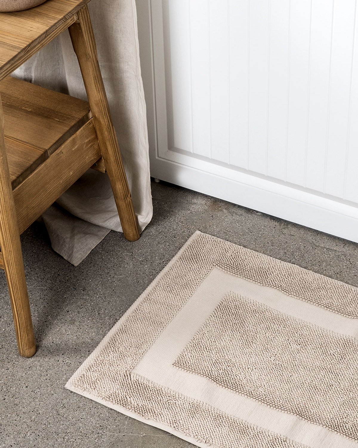 Kaia Aegean Cotton Bath Mat in Whispering Sands, placed in front of a bathroom door, showcasing its soft texture, warm hue, and luxurious quality