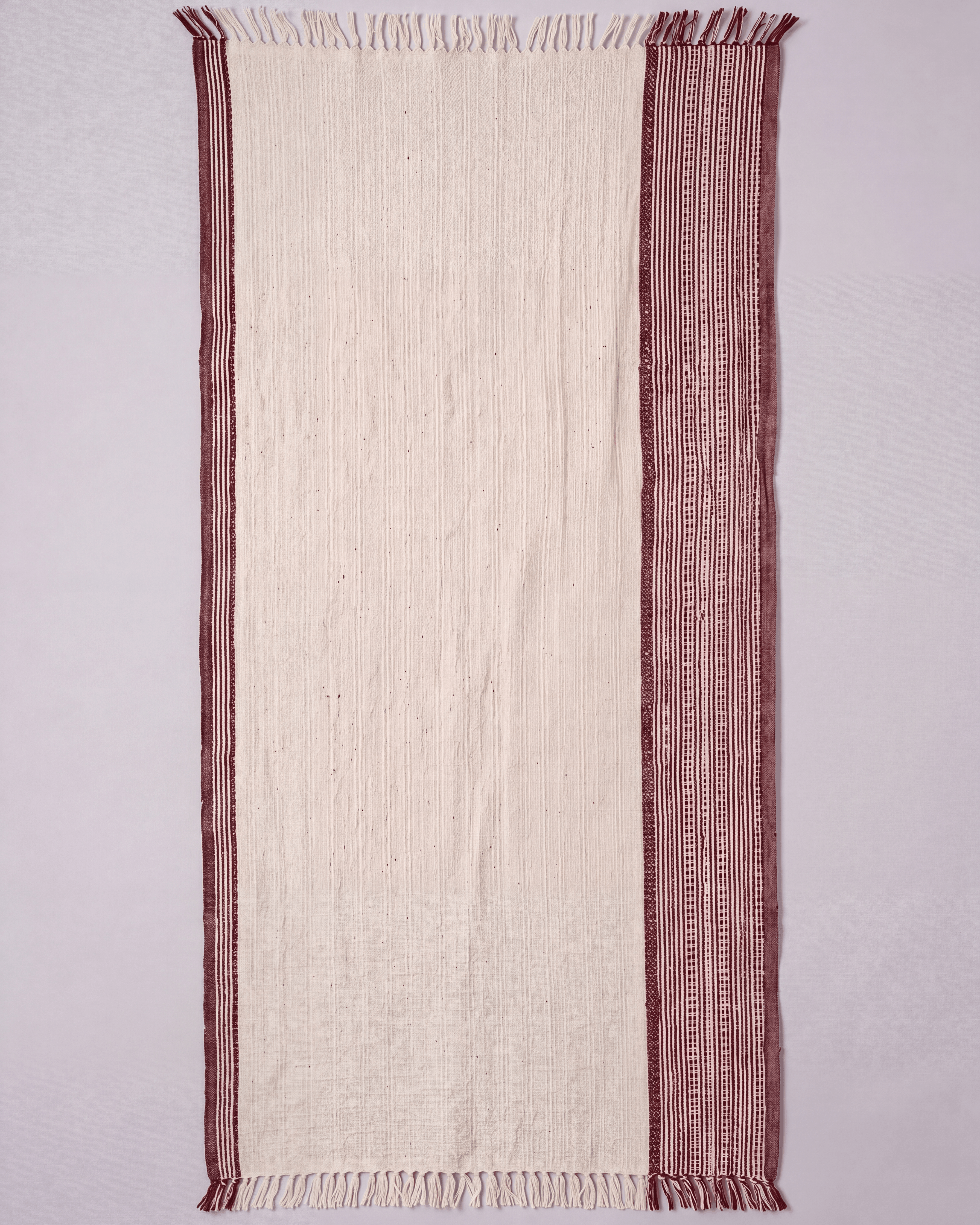 A rectangular cream-colored woven towel with fringed edges and a wide dusty rose patterned border, laid flat against a light gray background.