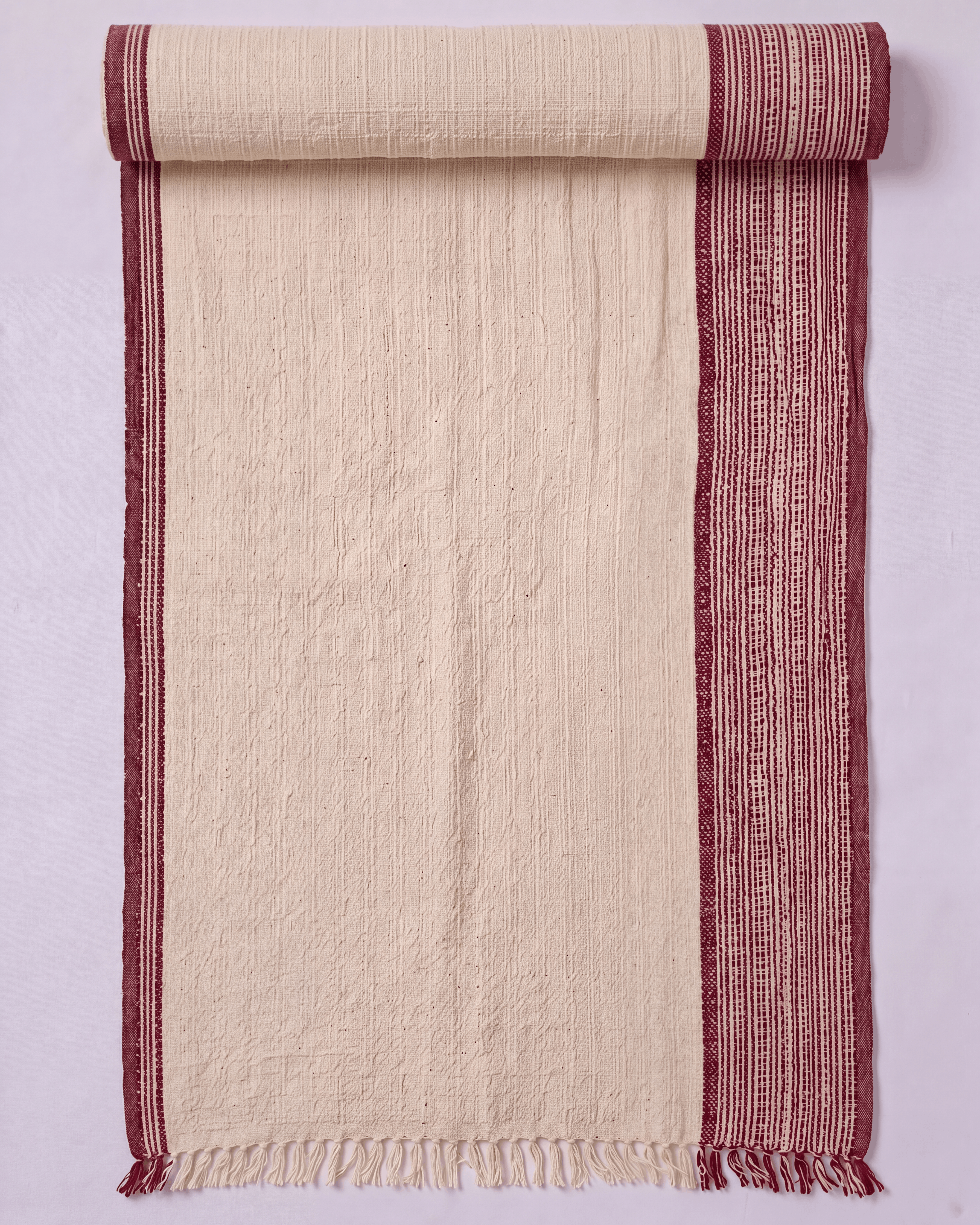 A cream-colored towel with rust-red borders, shown partially unrolled against a light lavender background.