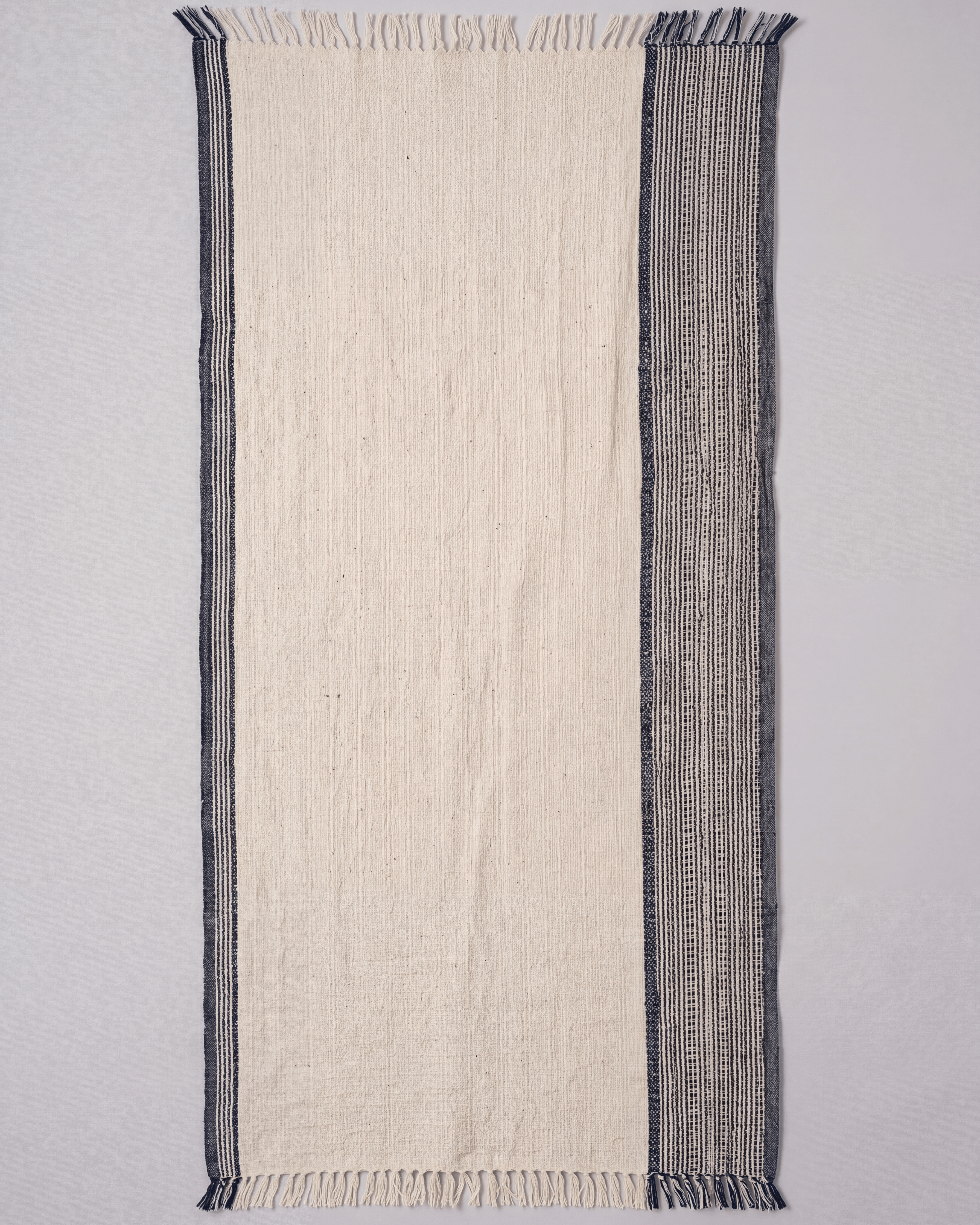 A rectangular woven towel featuring a wide off-white center panel, thin dark navy borders on the left, and a wider dark blue vertical patterned band on the right, finished with short fringe on both ends.