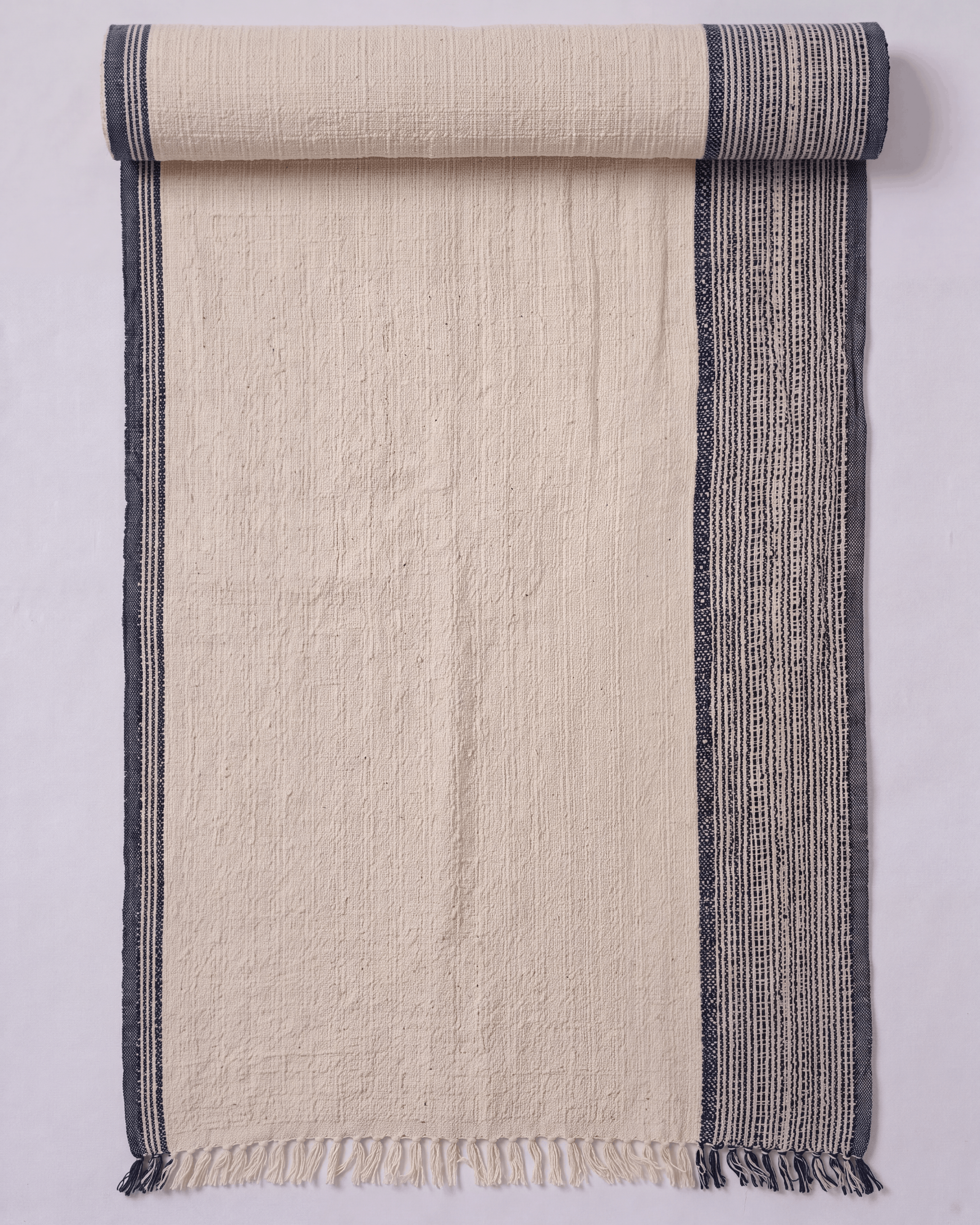A cream-colored woven towel shown partially rolled at the top. It features a textured off-white center with a dark gray patterned border on the right side and a solid dark gray stripe on the left edge, finished with a fringed hem.