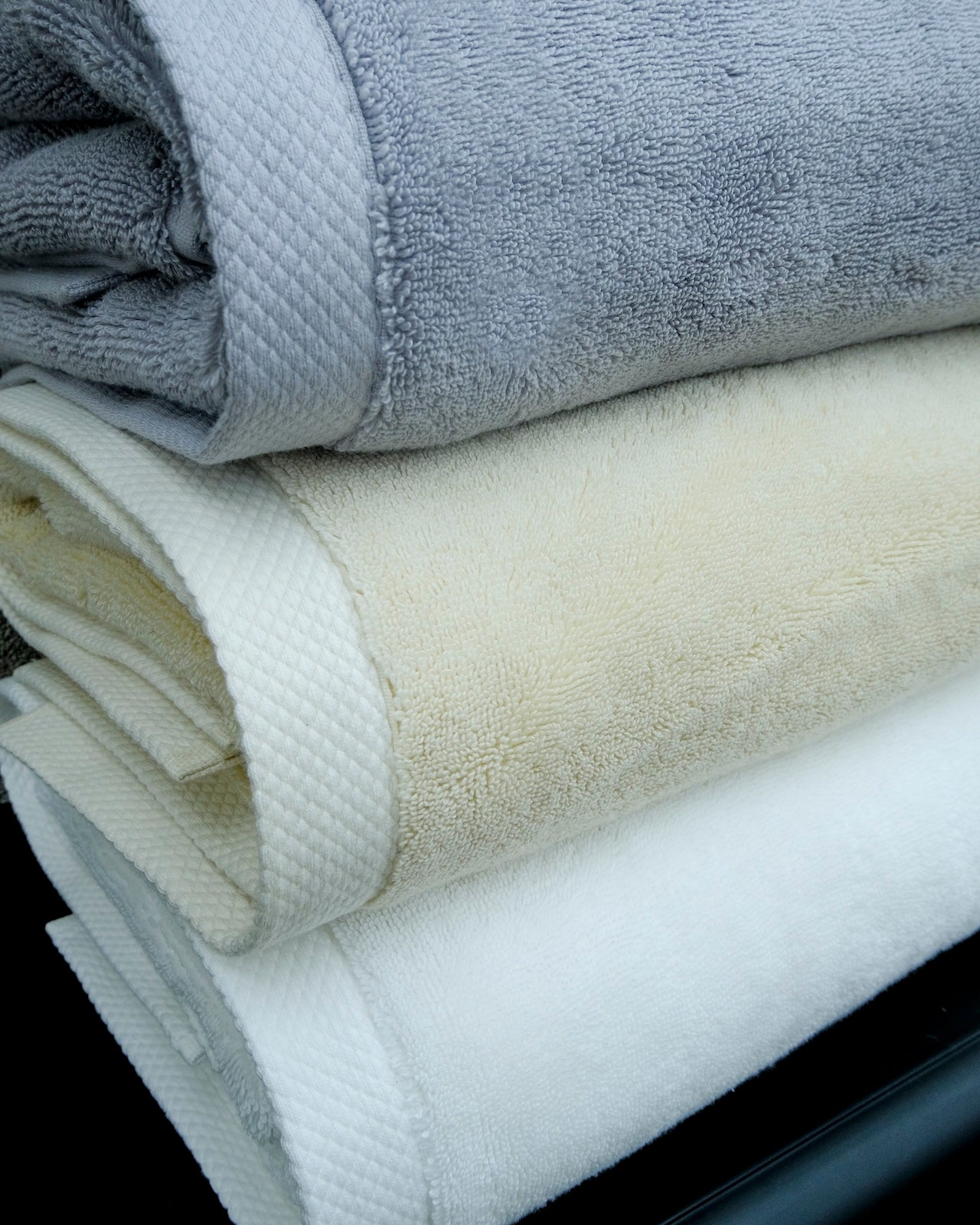 Close-up of a stack of three luxury organic cotton bath towels in white, cream, and grey from Minoa Home, displayed on a marble background