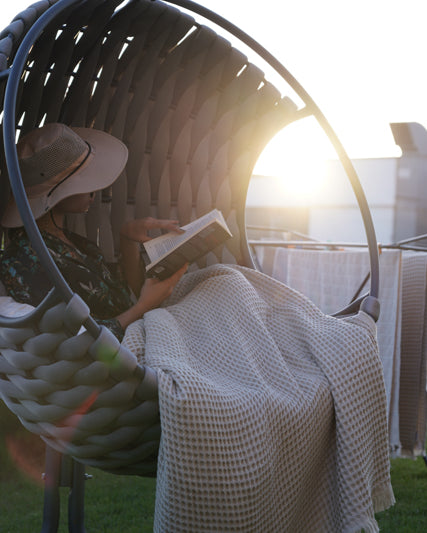 Woman reading on circular chair, legs covered by beige Organic Aegean Seren Waffle Large Throw, 400g organic cotton sheets.