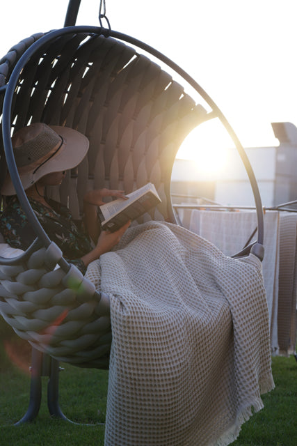 Woman reading on circular chair, legs covered by beige Organic Aegean Seren Waffle Large Throw, 400g organic cotton sheets.