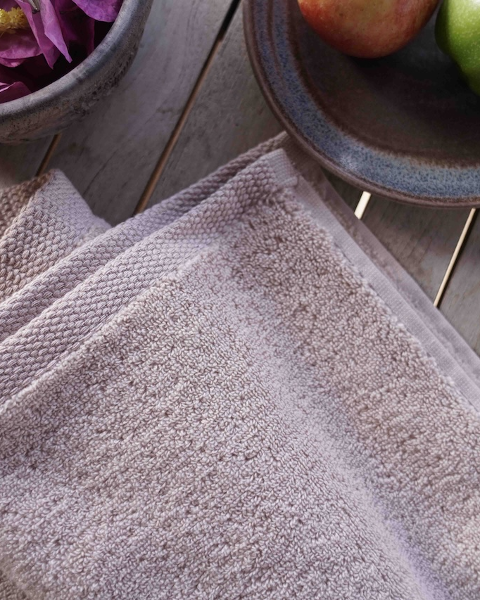 Stack of gray towels on a wooden surface with a bowl of flowers and apples in the background.