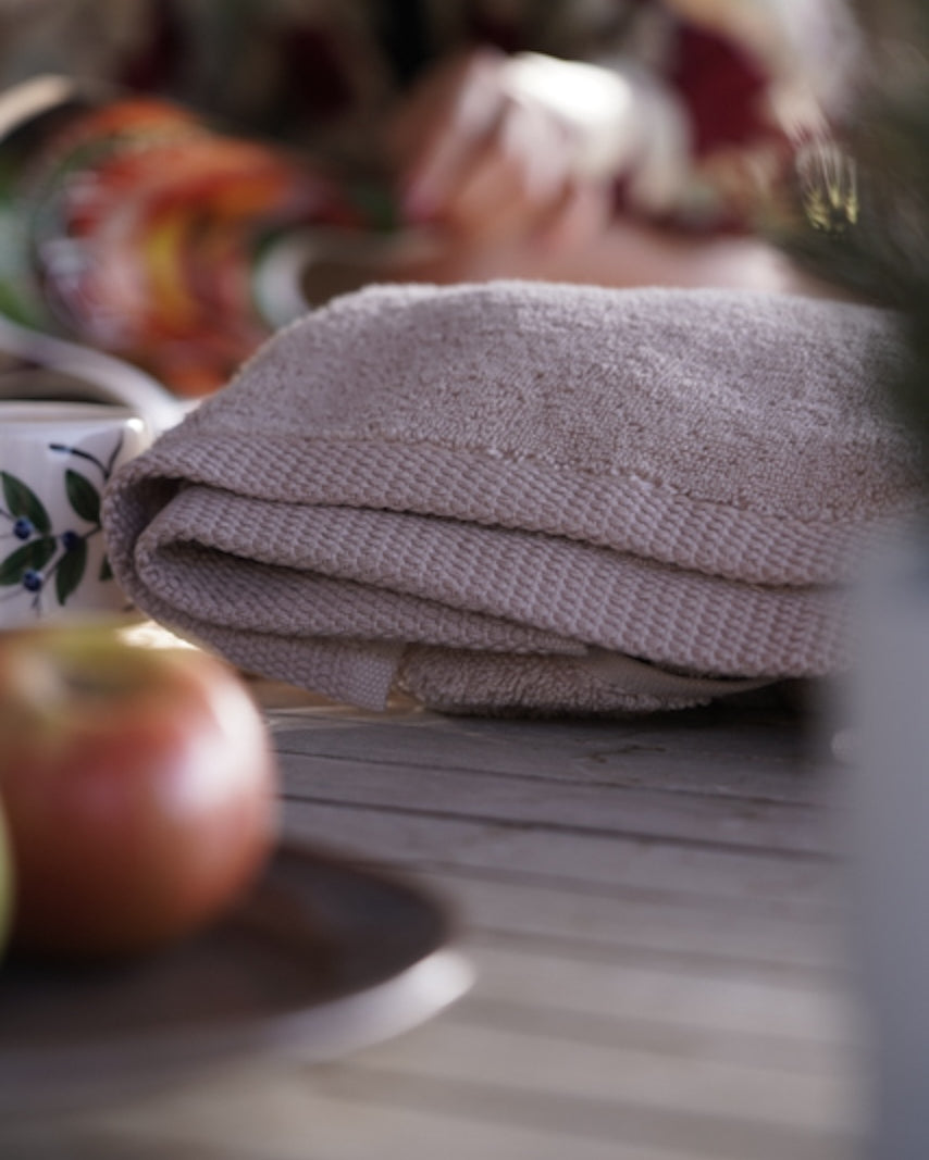 Folded beige towel on a wooden surface with an apple and a cup in the foreground.