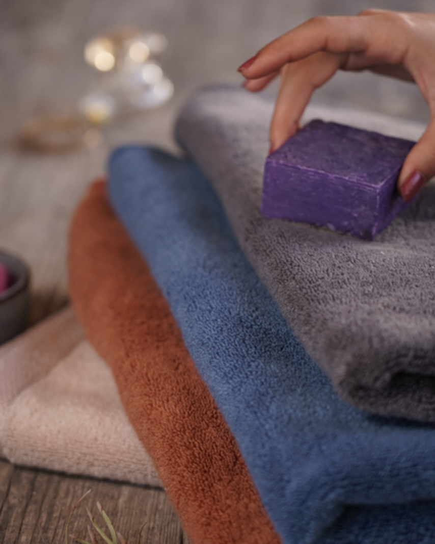 Hand holding a purple bar of soap over stacked colorful towels with a blurred background.