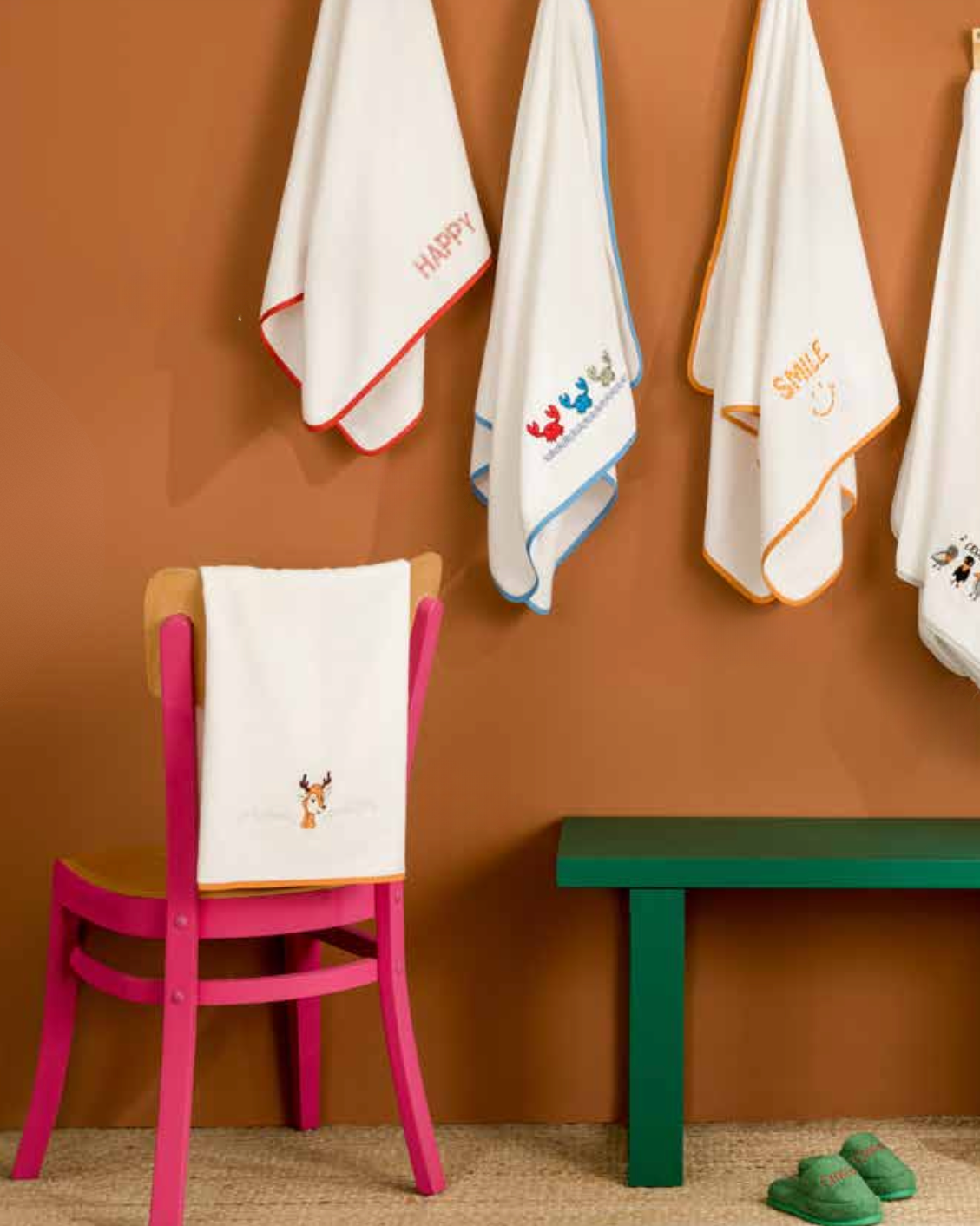 White towels with embroidered designs hanging on a brown wall, with a pink chair and green bench below.