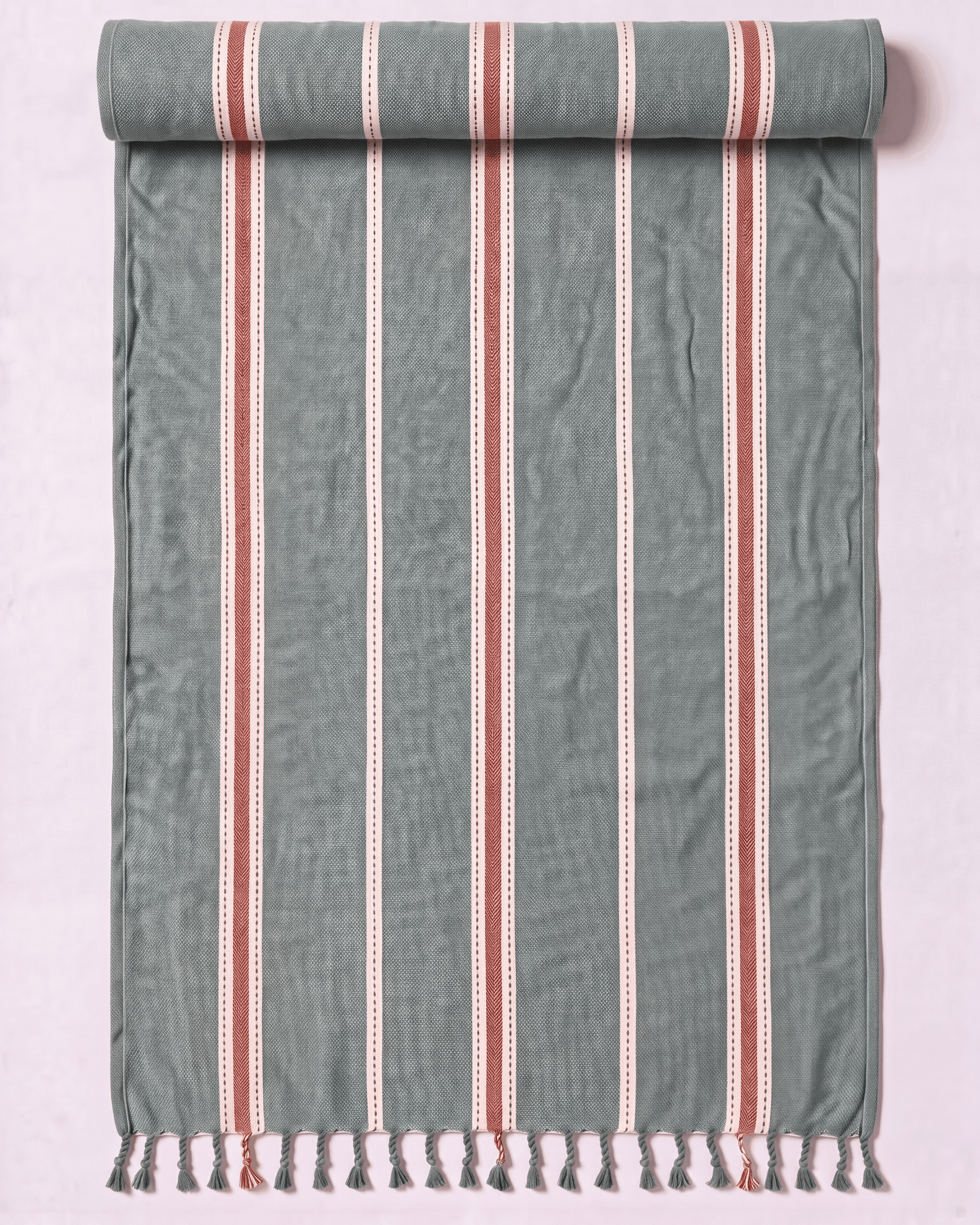 A rolled-out cotton Turkish towel with vertical stripes in sage green, terracotta red, and white, finished with a fringed tassel edge, shown against a light pink background.