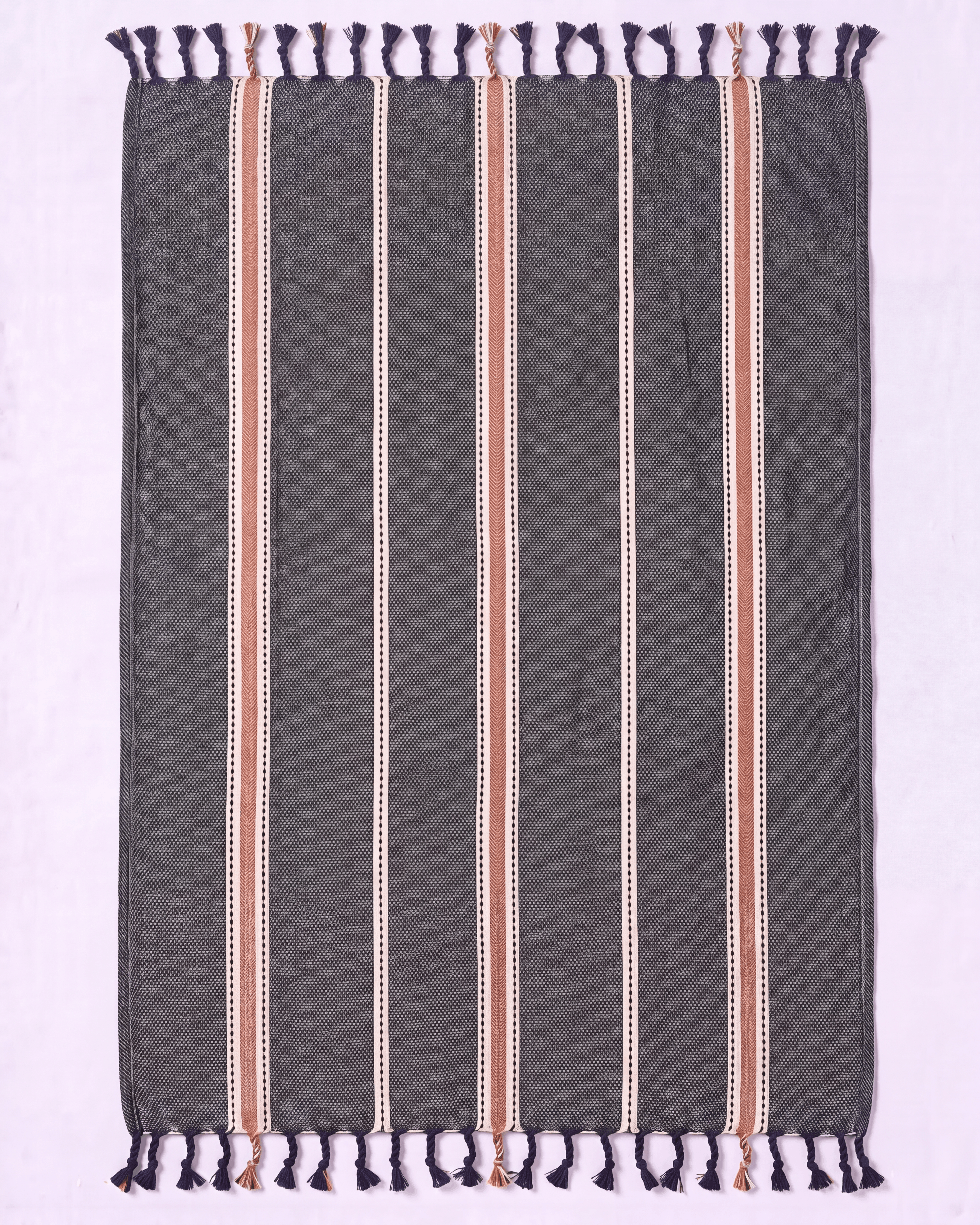 A rectangular woven towel with dark gray base, vertical stripes in beige, white, and muted pink, and dark purple fringe tassels on the top and bottom, laid flat on a light lavender background.