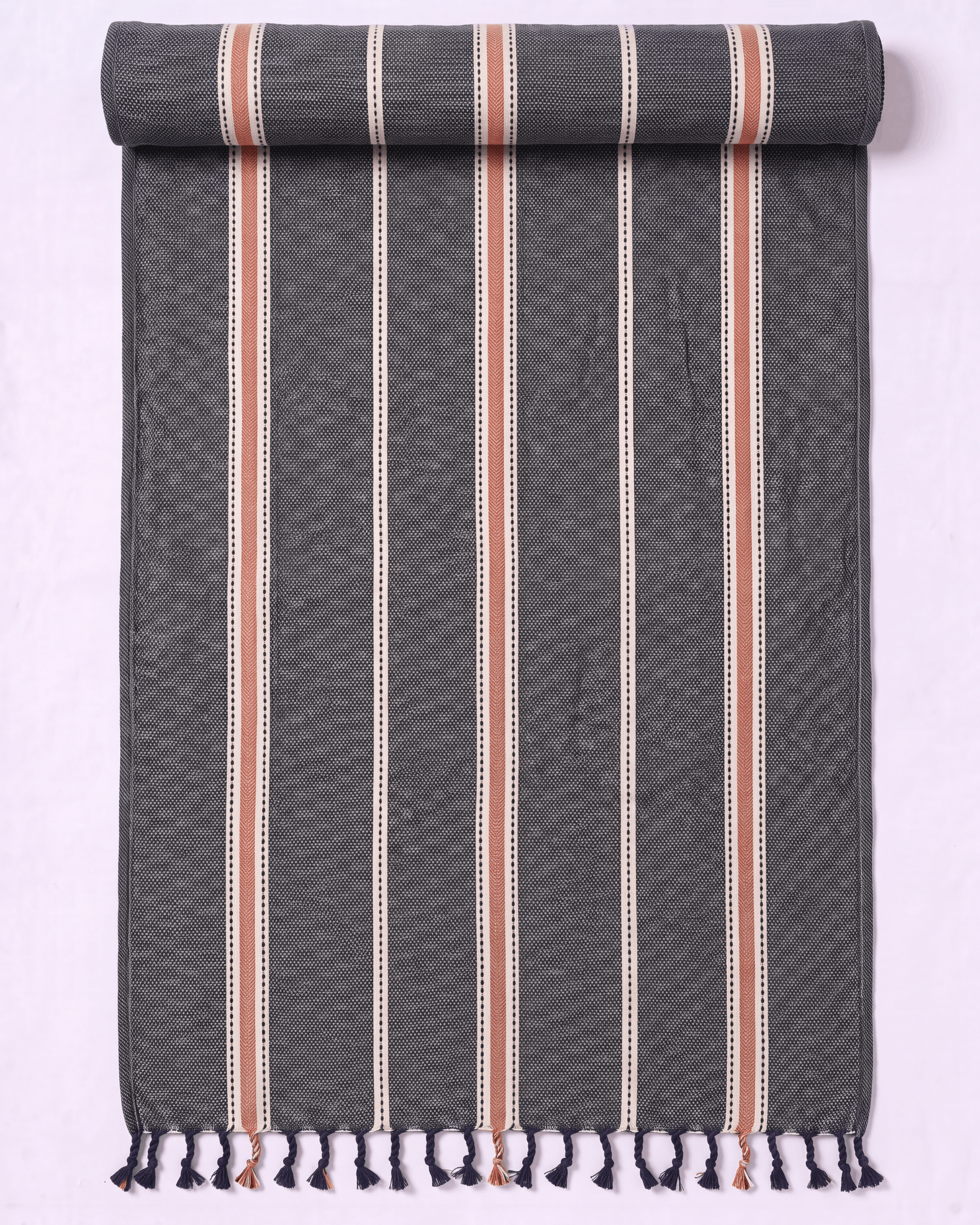 A dark gray woven towel with vertical stripes in cream, terracotta, and white, finished with fringe tassels at the bottom, shown partially rolled on a light lavender background.