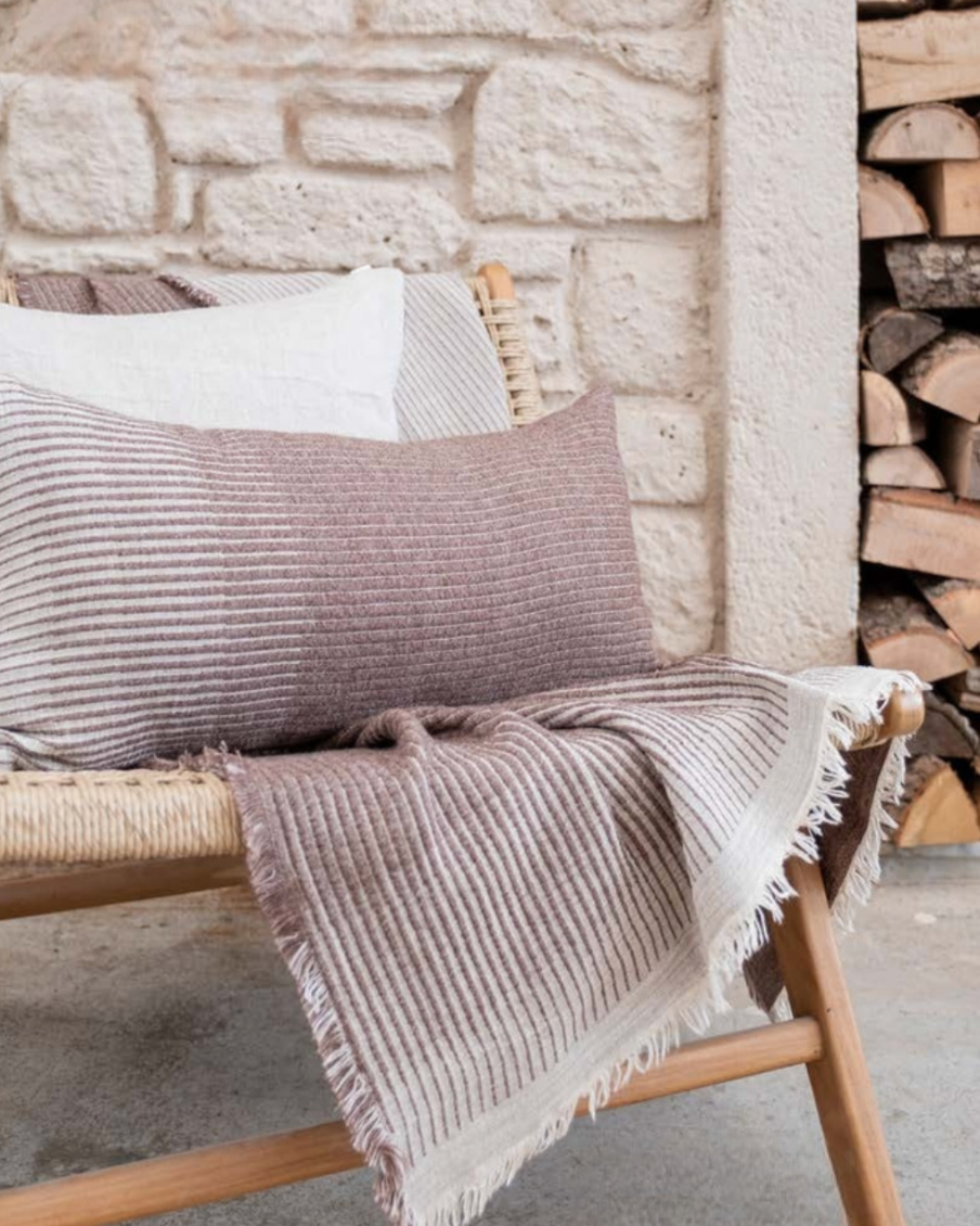Minoa Amaya collection featuring a striped cushion and bed cover in soft earthy tones on a woven bench, elegant sustainable home decor with artisanal detail.