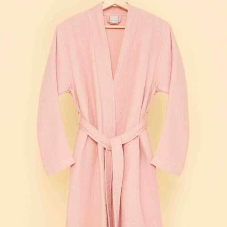 Blush pink bathrobe with belt tie hanging on wooden hanger against soft pastel background — minimalist eco-luxury loungewear aesthetic.