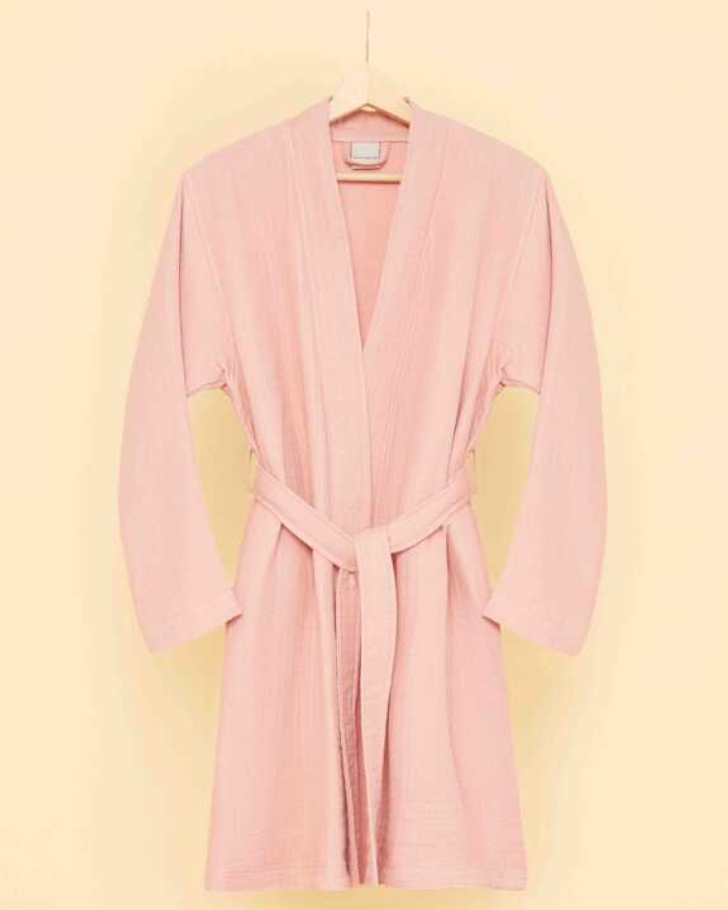 Blush pink bathrobe with belt tie hanging on wooden hanger against soft pastel background — minimalist eco-luxury loungewear aesthetic.