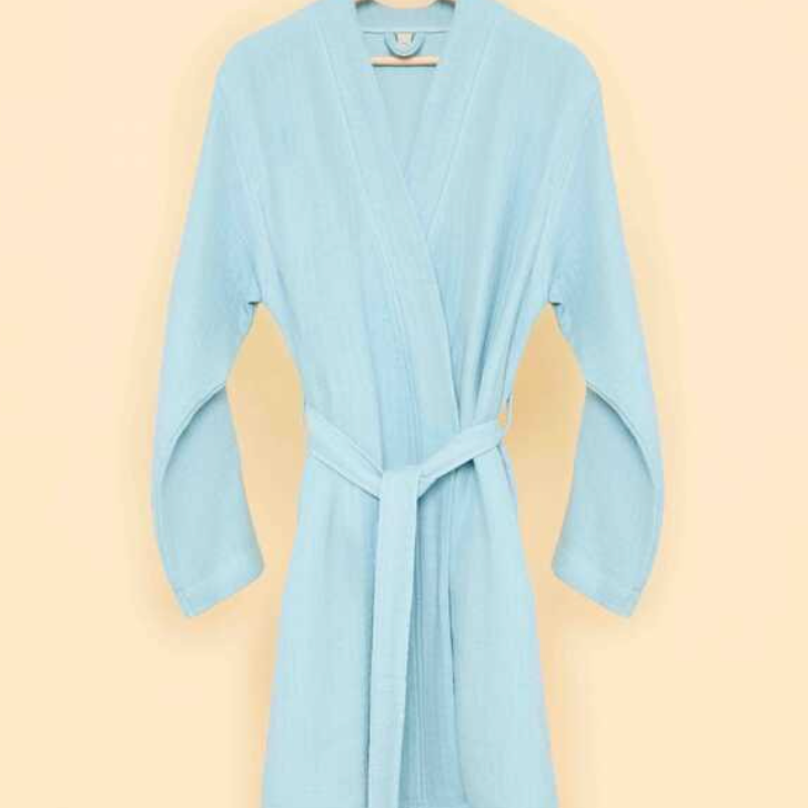 Light blue bathrobe with belt tie on wooden hanger against a soft beige background — minimalist sustainable loungewear aesthetic.