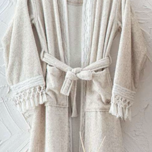 Beige textured bathrobe with tassel trim hanging against a white wall, folded towel and diffuser in foreground — luxury eco-conscious home textile style.