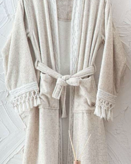 Beige textured bathrobe with tassel trim hanging against a white wall, folded towel and diffuser in foreground — luxury eco-conscious home textile style.