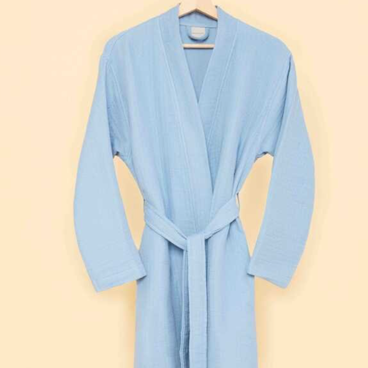 Sky blue bathrobe with waist tie hanging on wooden hanger against cream-colored wall — modern sustainable homewear with minimalist design.