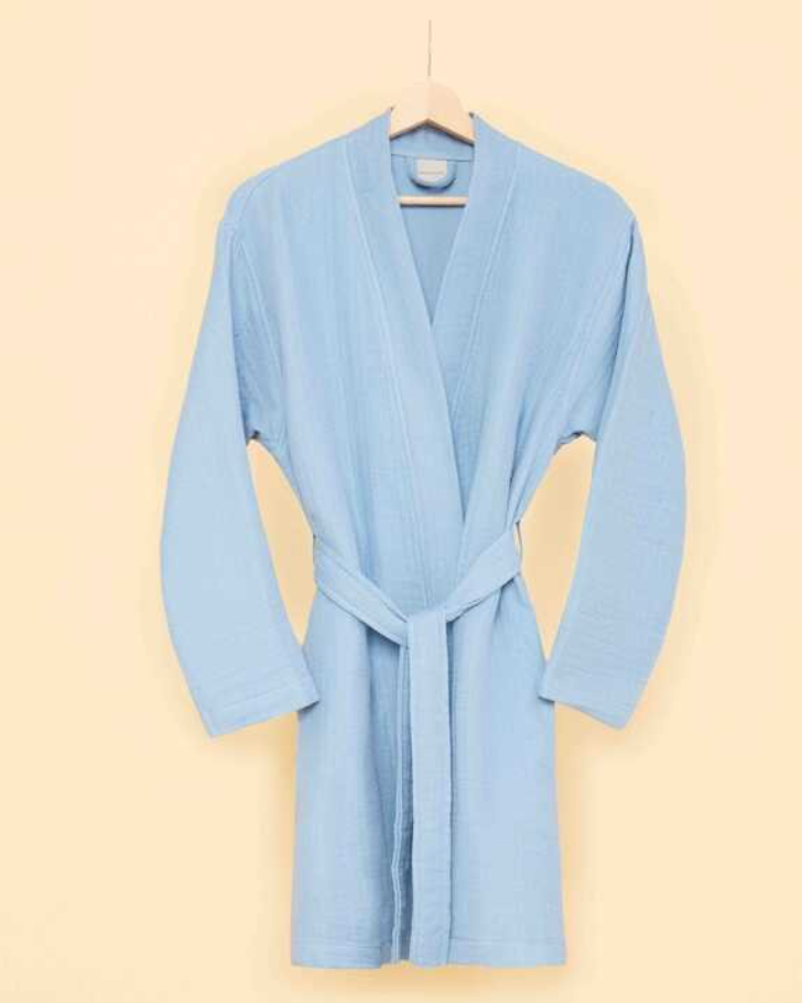 Sky blue bathrobe with waist tie hanging on wooden hanger against cream-colored wall — modern sustainable homewear with minimalist design.