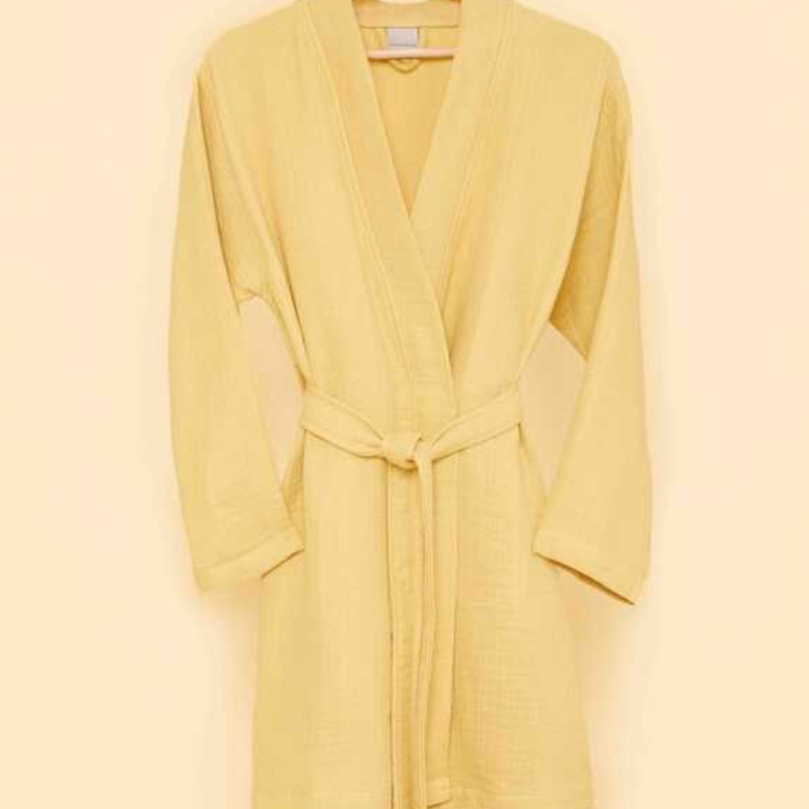 Soft yellow bathrobe with belt tie on wooden hanger against a warm beige wall — trendy eco-conscious loungewear for sustainable living.