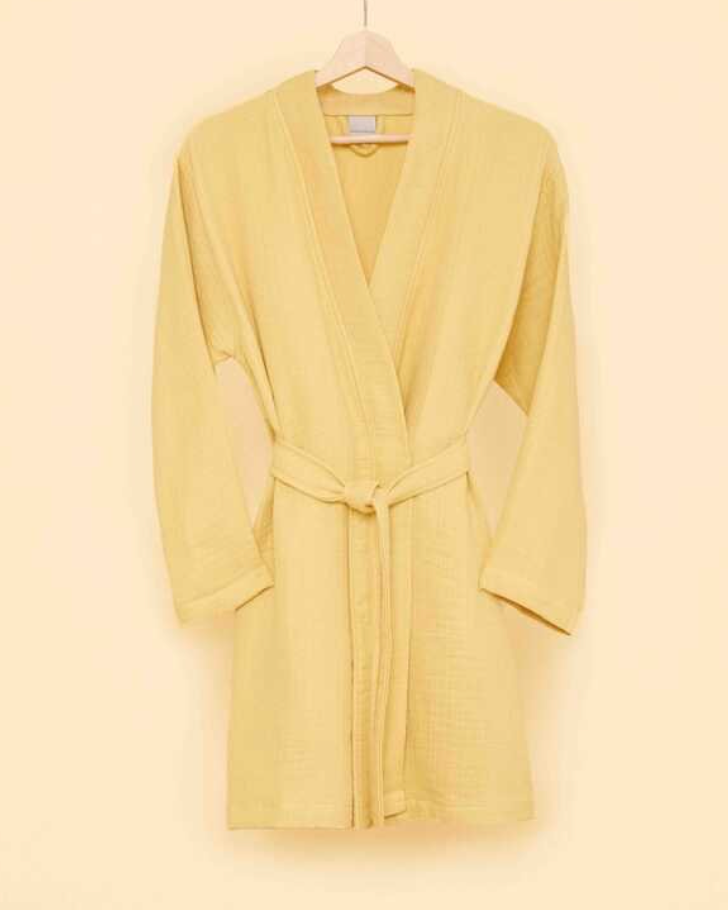 Soft yellow bathrobe with belt tie on wooden hanger against a warm beige wall — trendy eco-conscious loungewear for sustainable living.