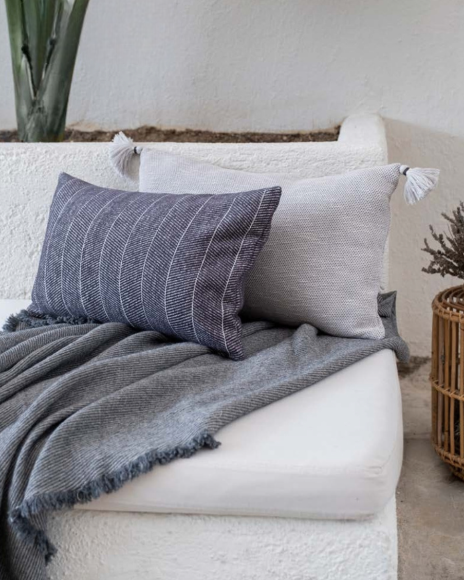 Bone-colored and navy striped pillows with tasseled corners styled on a textured sofa with a grey fringed throw — modern eco-luxury home decor.