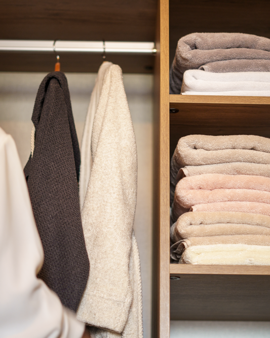 Luxury stacked bath towels in beige, blush, and cream tones neatly arranged in wooden closet shelves beside hanging robes and garments.