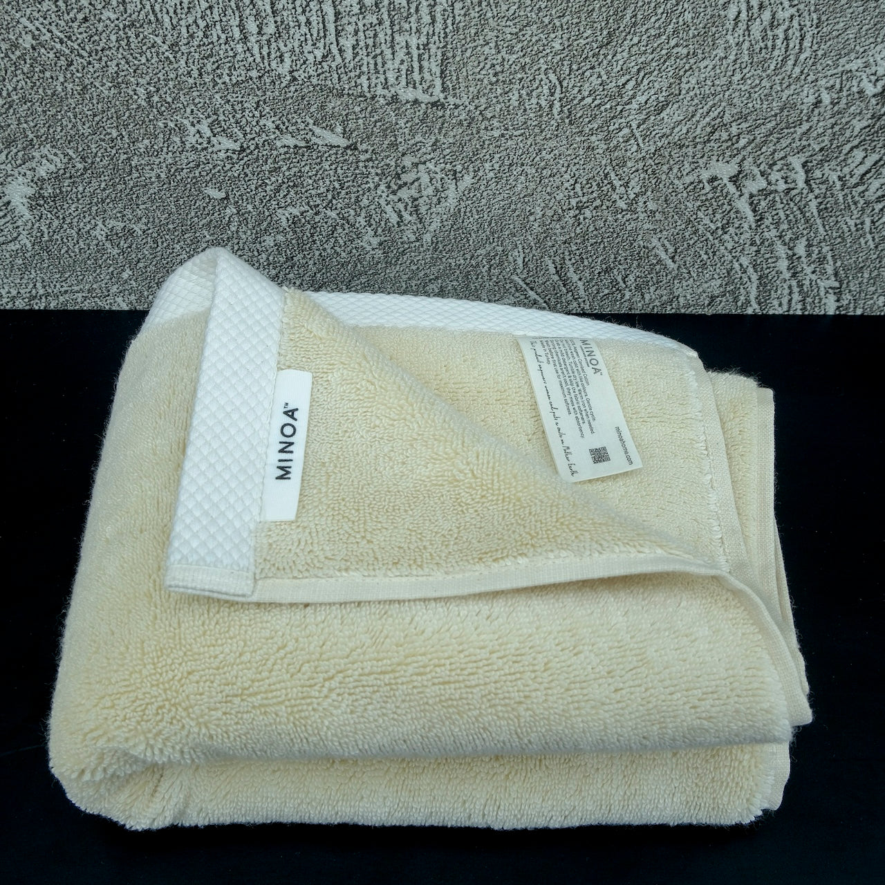 Folded Minoa Home cream-colored luxury bath towel with label visible, made from ultra-soft organic cotton, placed on a black surface with a textured background.