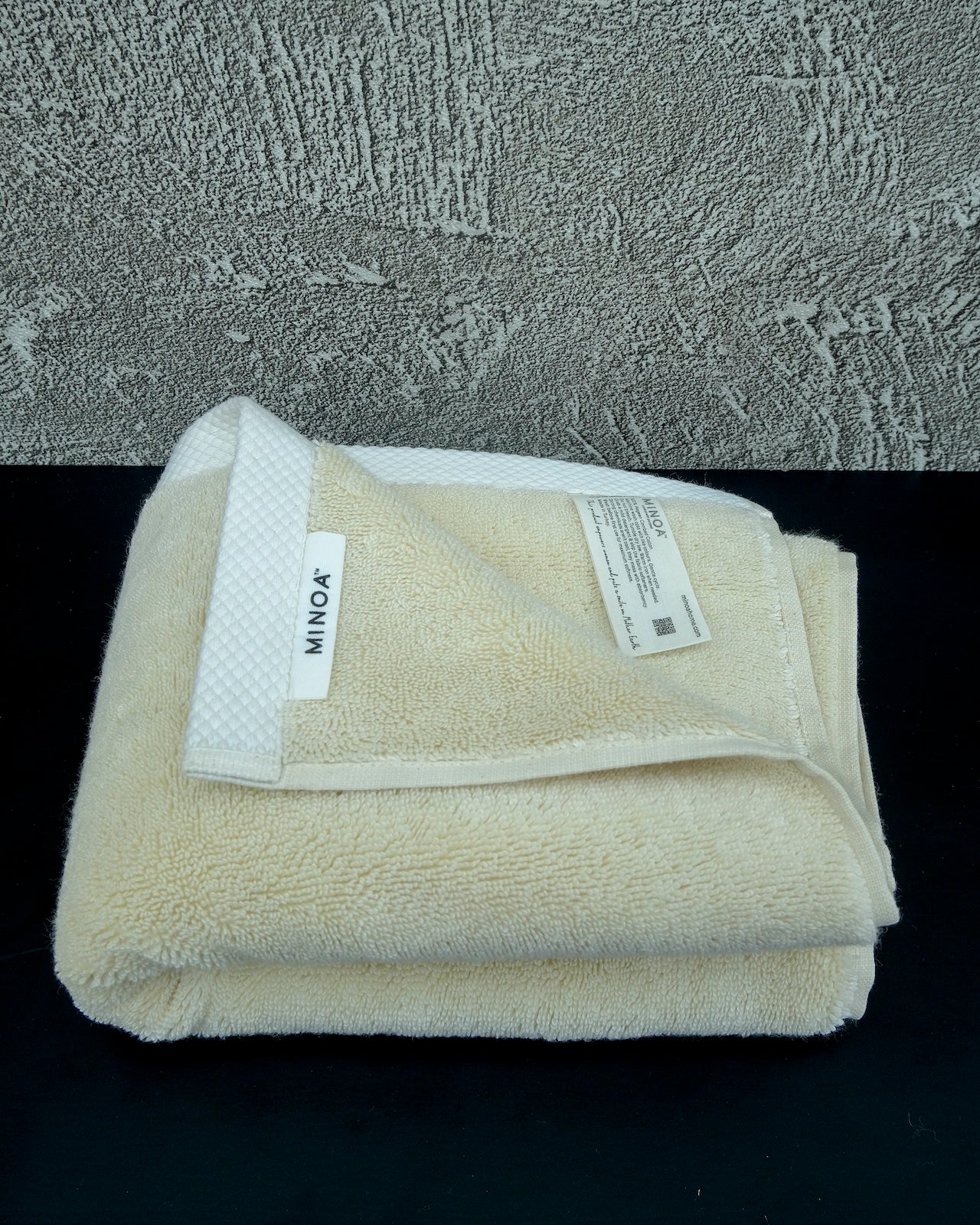 Folded Minoa Home cream-colored luxury bath towel with label visible, made from ultra-soft organic cotton, placed on a black surface with a textured background.