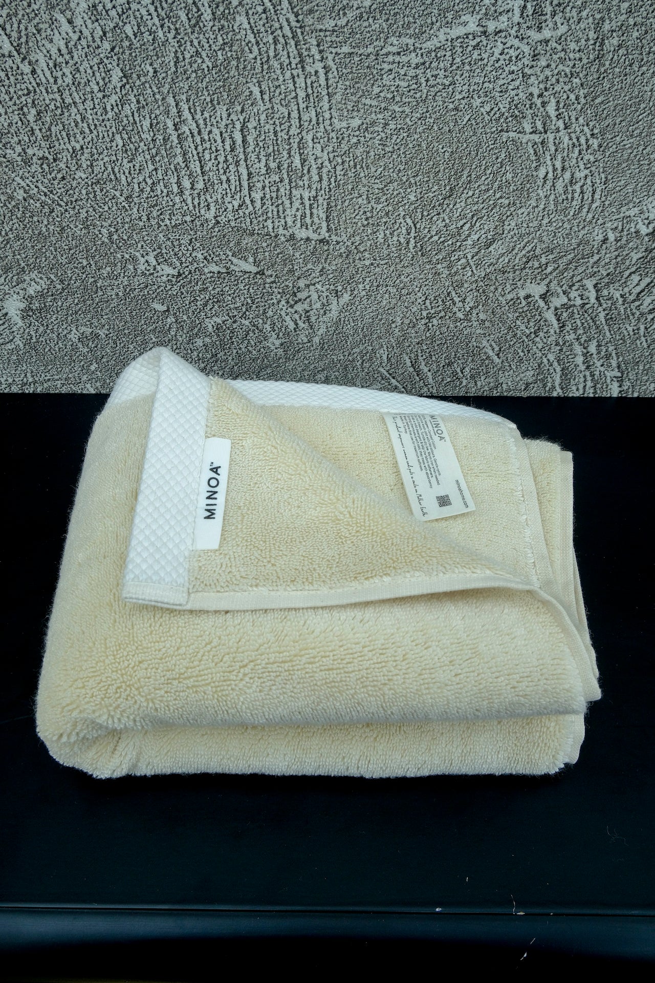 Folded Minoa Home cream-colored luxury bath towel with label visible, made from ultra-soft organic cotton, placed on a black surface with a textured background.