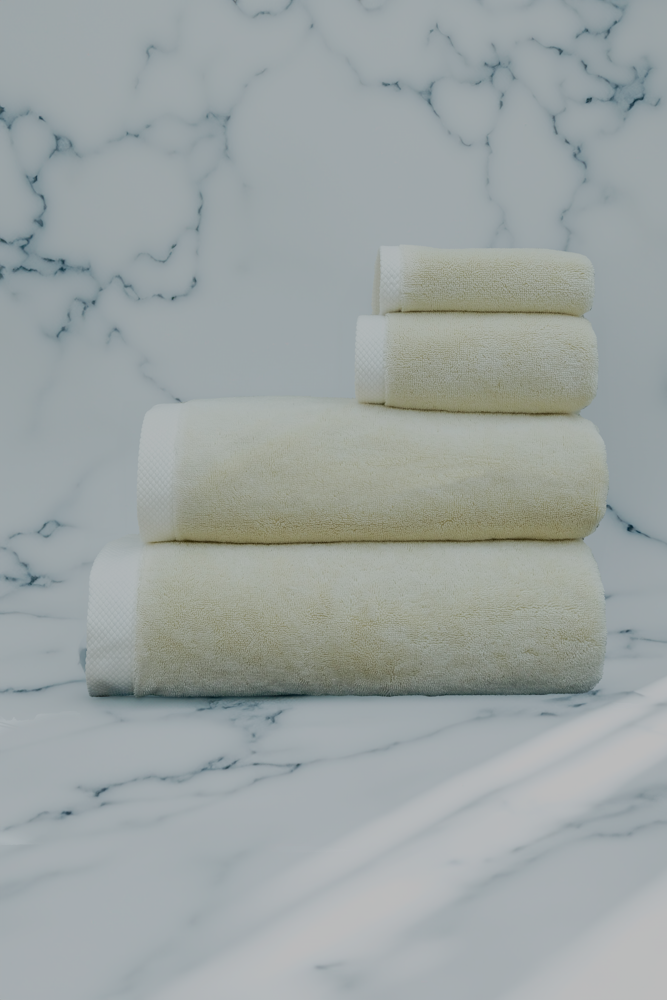 Cream-colored set of luxury organic bath towels from Minoa Home, including bath, hand, and face towels, neatly arranged on a white marble background
