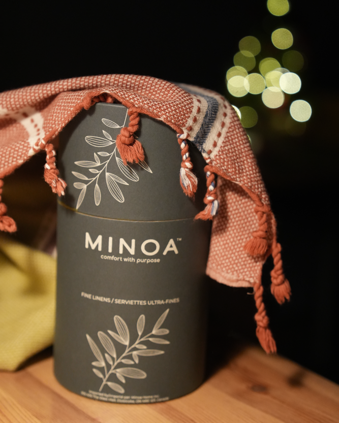Minoa Home fine linens gift packaging with a red tasseled serviette draped over a luxurious dark grey box, festive blurred lights in background.