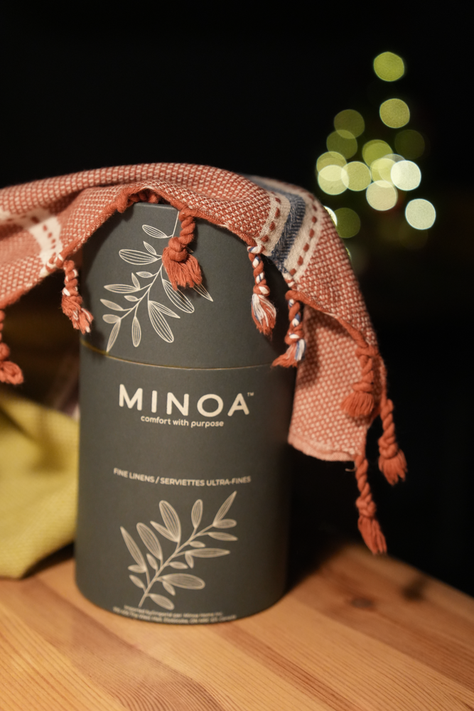 Minoa Home fine linens gift packaging with a red tasseled serviette draped over a luxurious dark grey box, festive blurred lights in background.