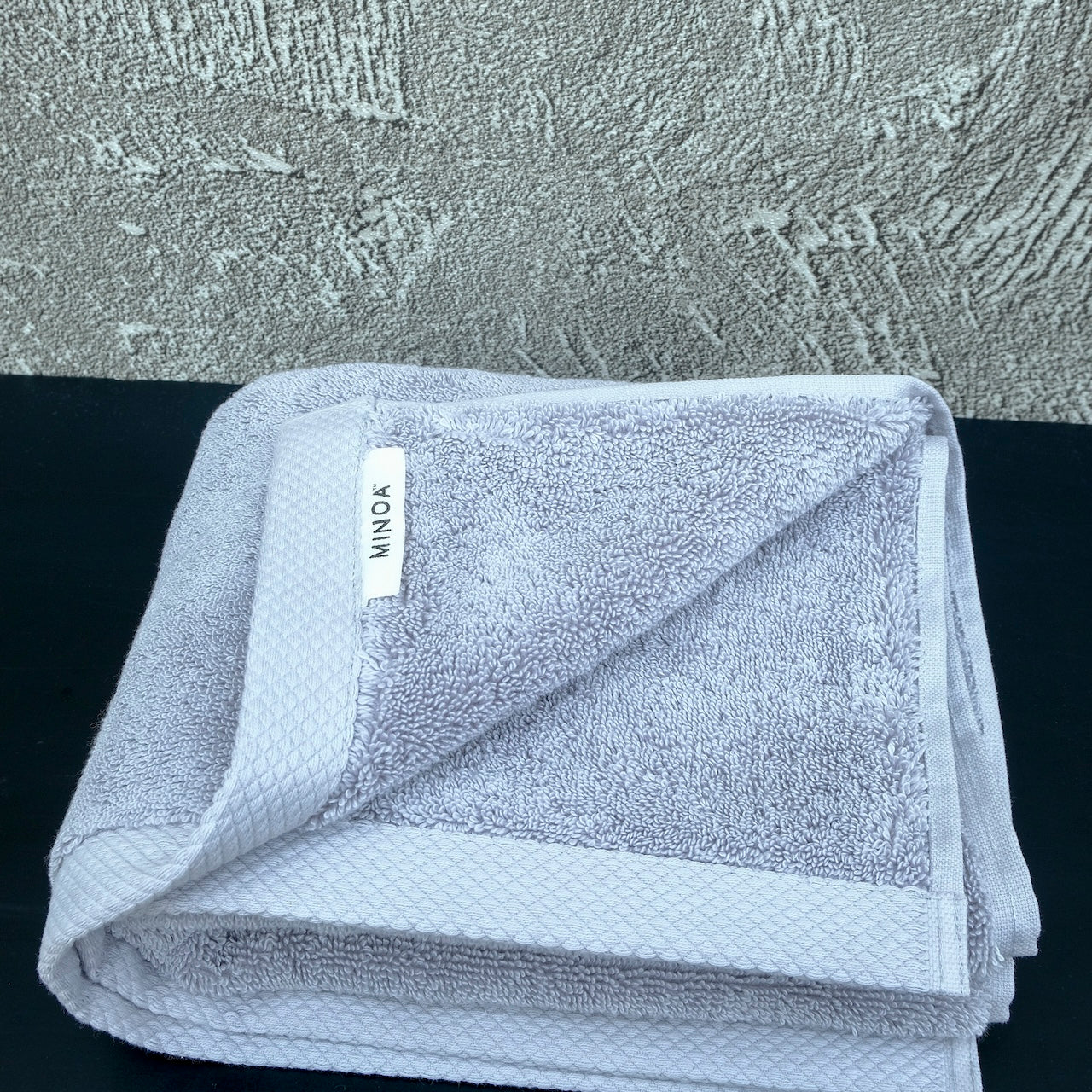 Soft grey luxury cotton towel by Minoa Home, neatly folded with a textured grey wall and black surface backdrop, showcasing plush and elegant bath linen.