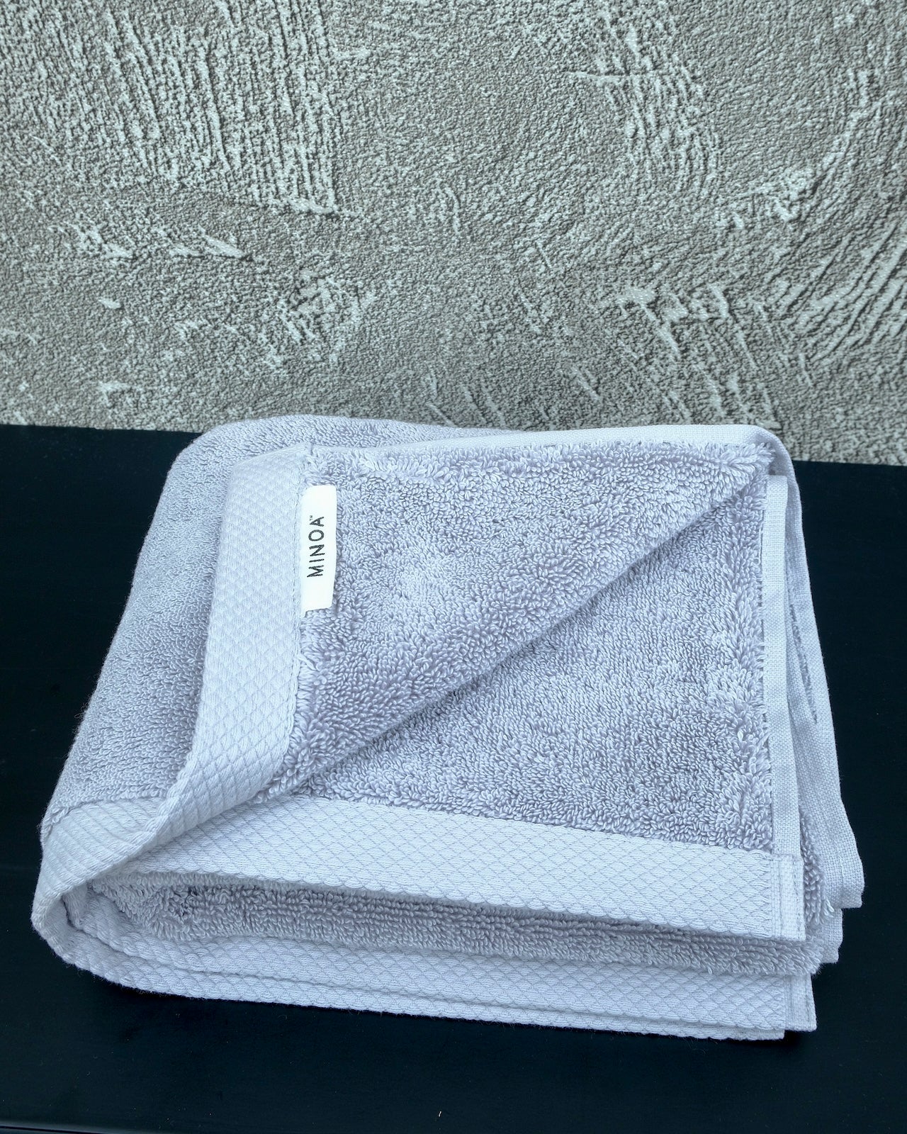 Soft grey luxury cotton towel by Minoa Home, neatly folded with a textured grey wall and black surface backdrop, showcasing plush and elegant bath linen.