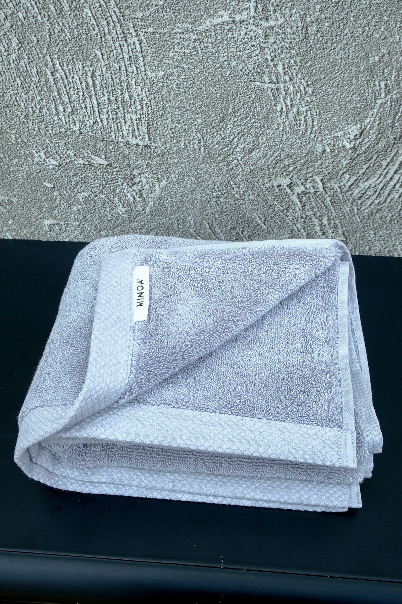 Soft grey luxury cotton towel by Minoa Home, neatly folded with a textured grey wall and black surface backdrop, showcasing plush and elegant bath linen.