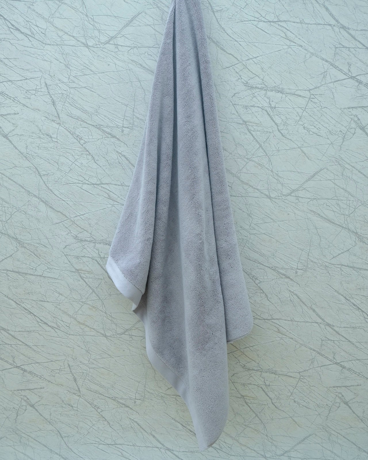 Soft grey Minoa Home organic cotton hand towel hanging on a marble-patterned wall, showcasing minimalist bathroom decor.