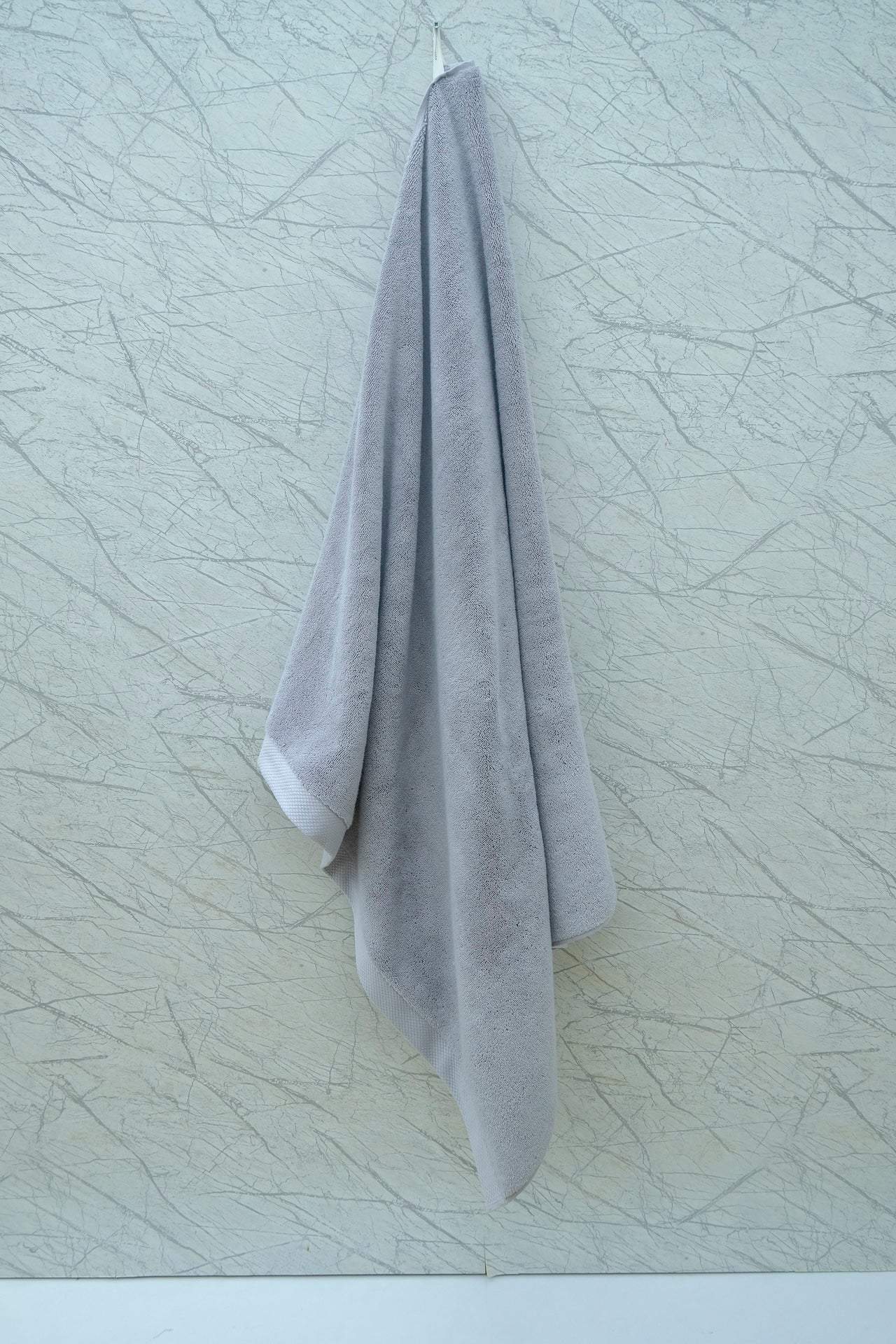 Soft grey Minoa Home organic cotton hand towel hanging on a marble-patterned wall, showcasing minimalist bathroom decor.