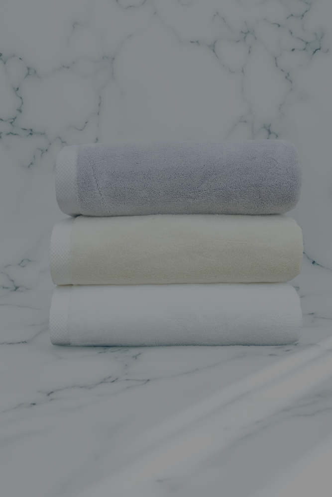Stack of three luxury organic cotton bath towels in white, cream, and grey from Minoa Home, displayed on a marble background