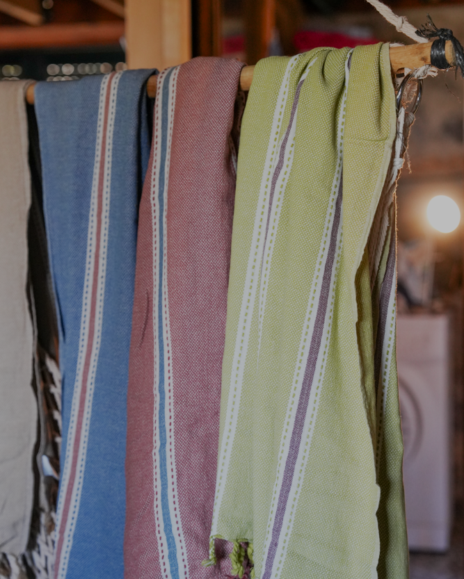 Minoa Home's multicolor peshtemal towels in blue, red, and green hues, handwoven with eco-friendly cotton and detailed white stripes, displayed on a rustic wooden rack.