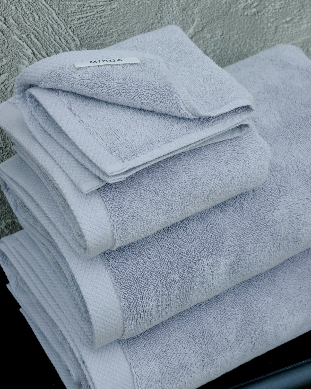 Stack of premium grey bath towels with visible MINOA tag, made from organic cotton, neatly folded against a textured wall.